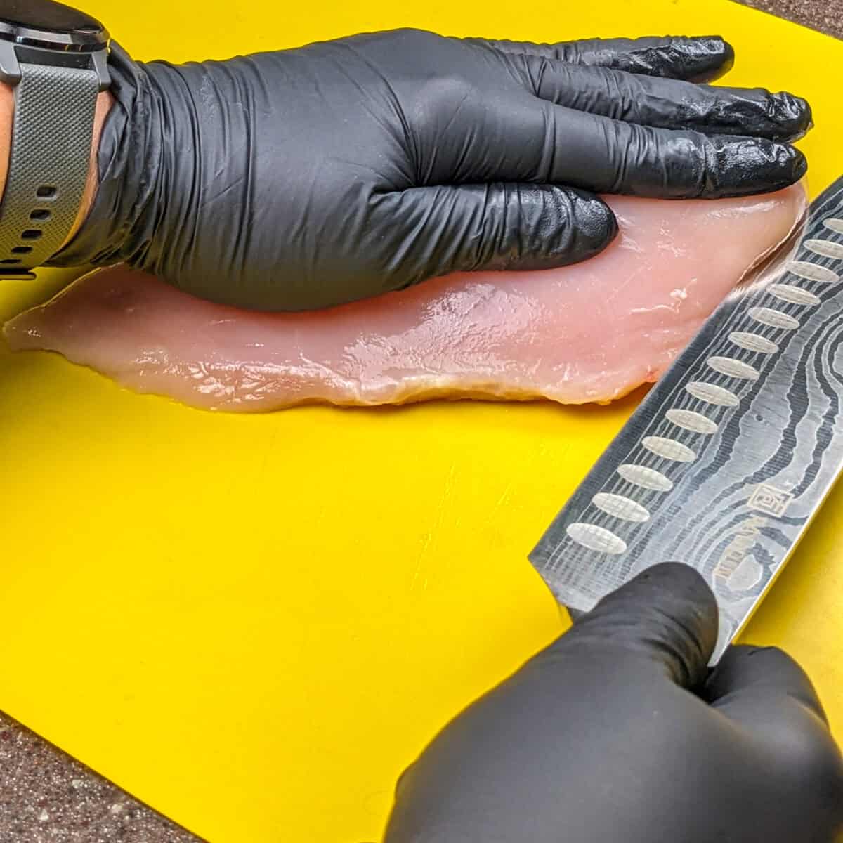 beginning step on how to butterfly a chicken breast with a knife at the tip side of the chicken breast on a cutting board.