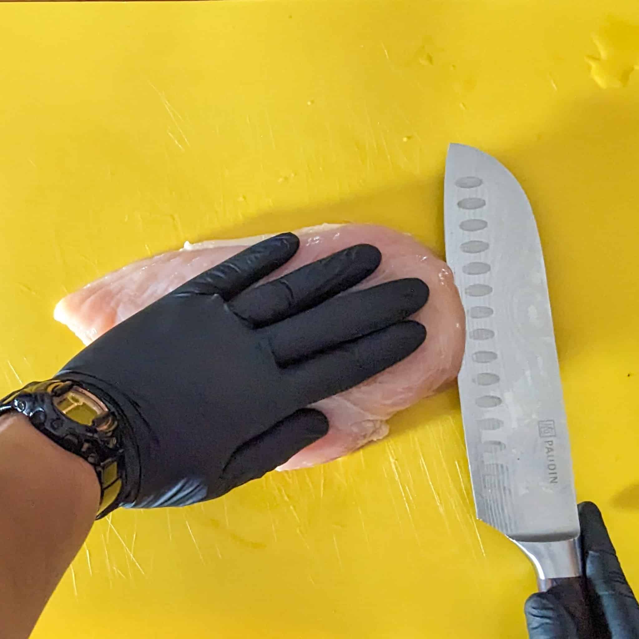 gloved hand on a chicken breast while the other hand slice it with a knife at the tip on a cutting board