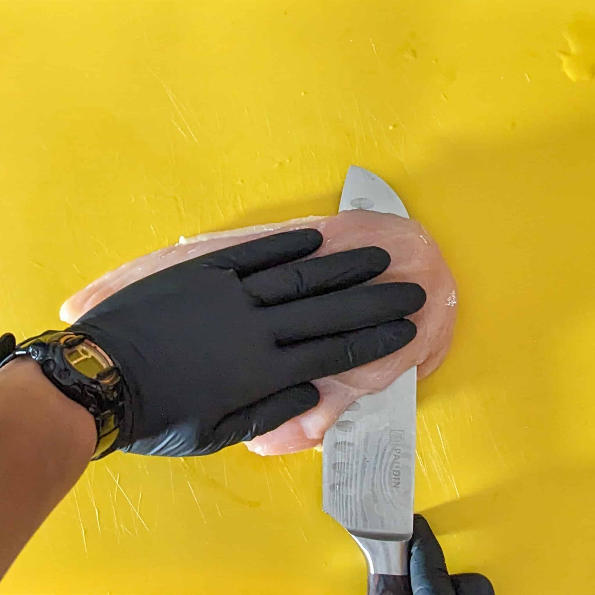 gloved hand on a chicken breast while the other hand slice it with a knife on a cutting board