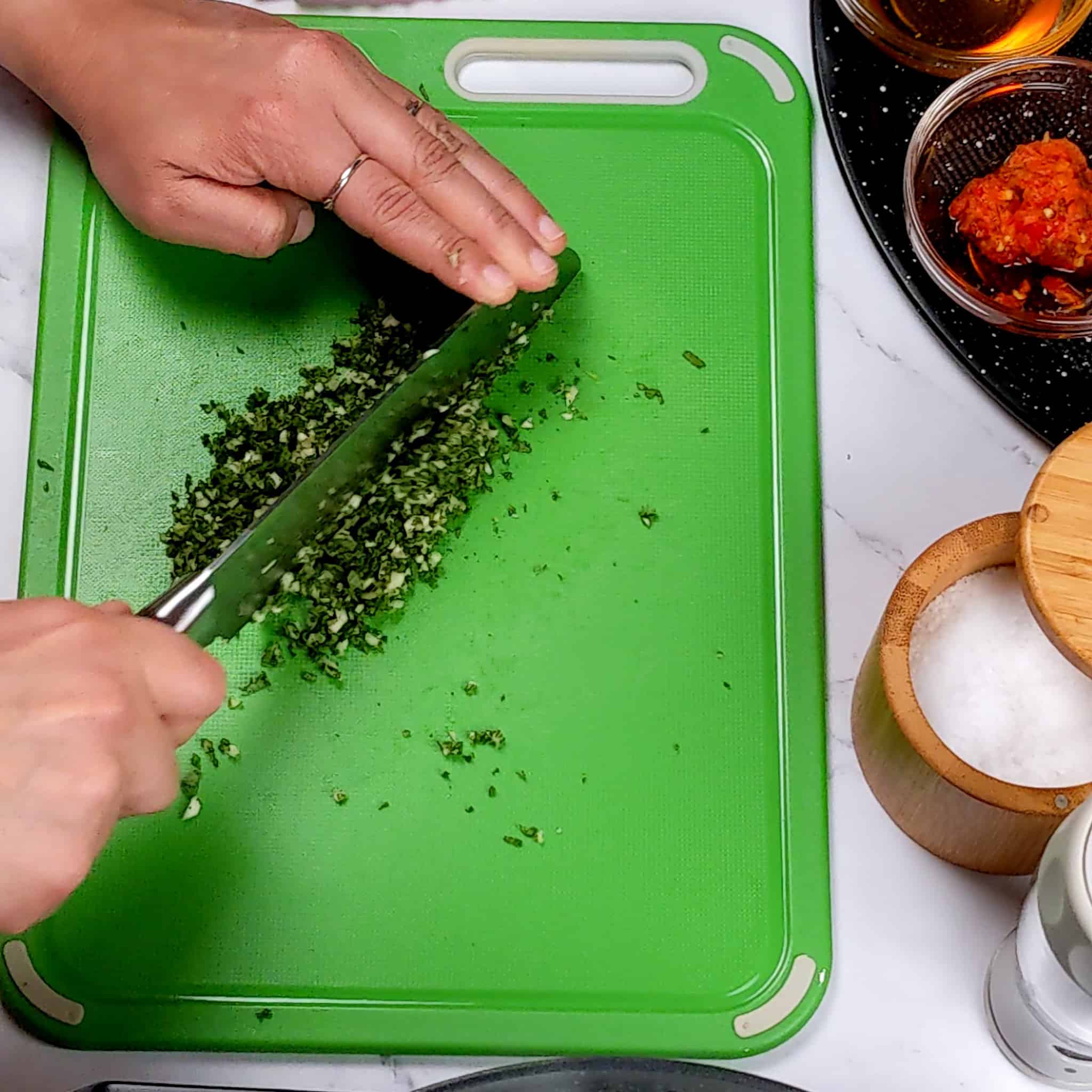 a knife finely chopping garlic and herbs on a plastic cutting board