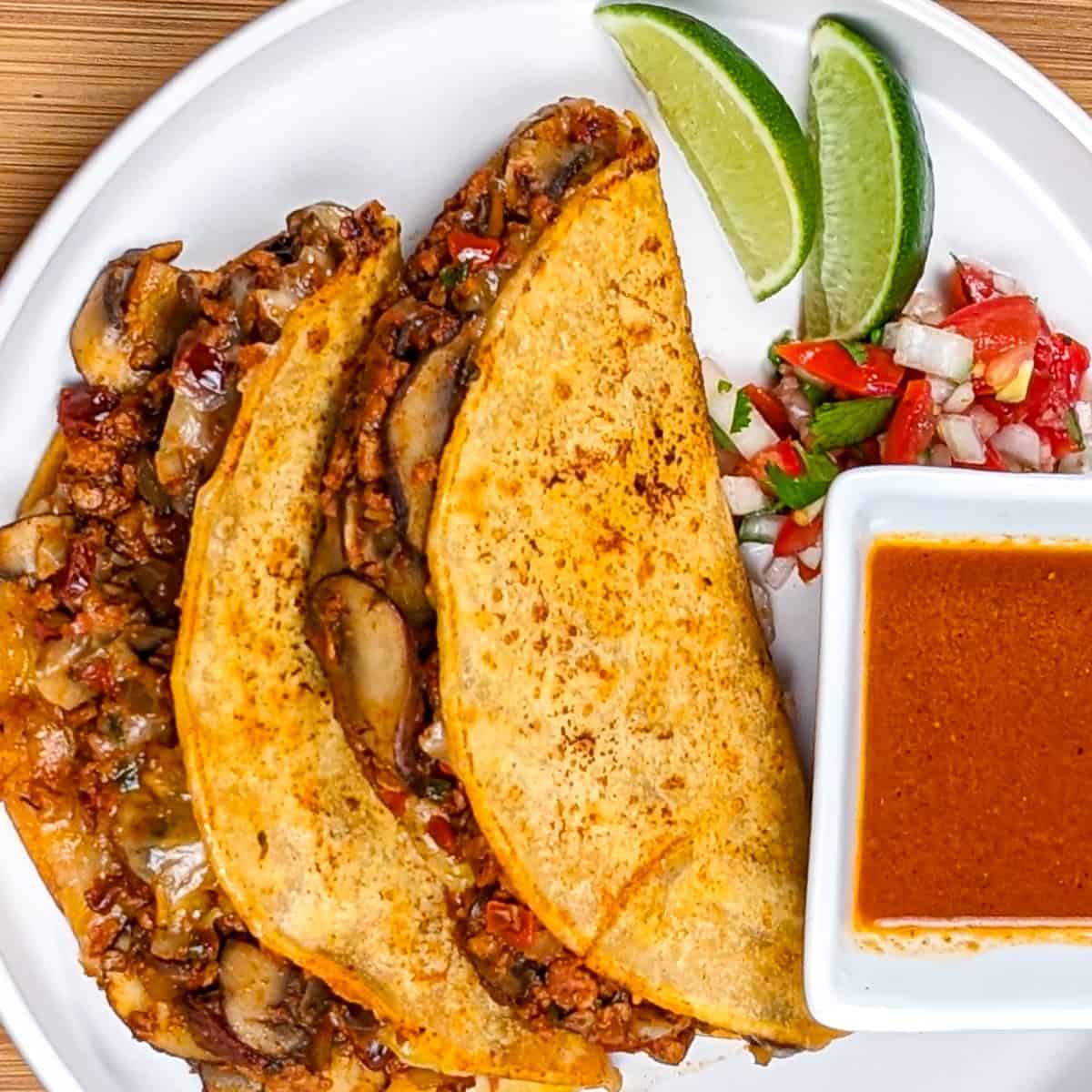 vegetarian mushroom stuffed birria tacos on a plate with consume, lime wedges and pico de gallo.