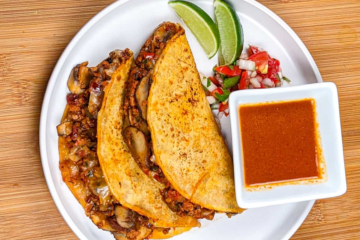 vegetarian mushroom stuffed birria tacos on a plate with consume, lime wedges and pico de gallo.