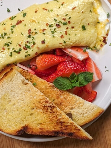Whipped Cottage Cheese stuffed French Omelet on a round white plate with sliced strawberries and toasted sourdough bread
