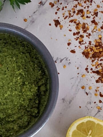 zhoug sauce on marble surface with a halved lemon, scattered red pepper chili flakes and parsley sprigs in the background