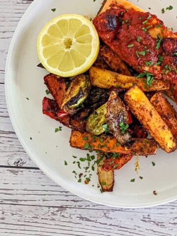 Roasted harissa salmon with paprika potatoes and brussel sprouts with half a lemon on a round white plate with parsley sprinkled on top.