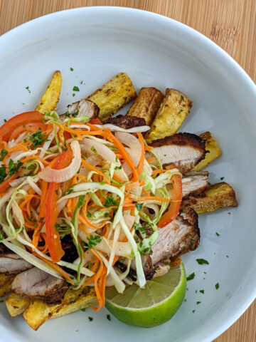 Plantain fries topped with sliced spiced chicken and a citrus pepper cabbage slaw in a white bowl.