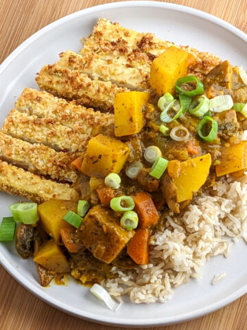 Breaded sliced tofu and brown rice topped with curried potatoes, carrots and onions on a white round plate.