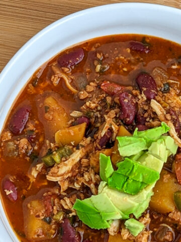 chili in a bowl with shredded chicken, butternutsquqsh, kidney beans, with diced avocado spooned on top in a white wide rim bowl