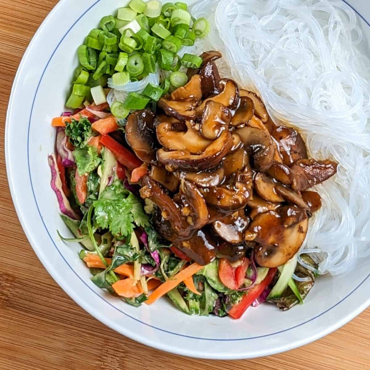 Hoisin mushrooms on top of vegetables in peanut dressing, sliced scallions and vermicelli noodles in a white and blue bowl