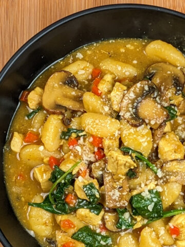 Gnocchi with small chicken chunks, brown sliced mushrooms, spinach and red diced bell pepper in a bowl of yellow curry sauce with sprinkled cheese on top