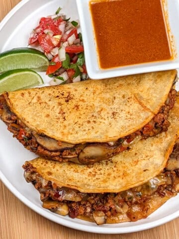 Two birria tacos stuffed with mushrooms and soy chorizo with two wedges of lime, a spoonful of pico de gallo and a white square ramekin of birria sauce all on a round white plate.