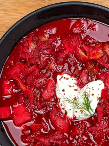 red borscht soup in a black bowl with sour cream in the shape of a heart on top sprinkled with aleppo pepper flakes and topped with a dill leaf.