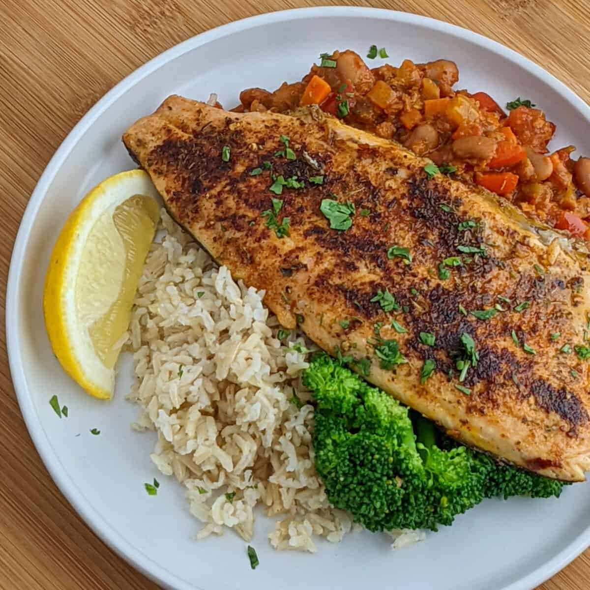 Blackened rainbow trout with spicy chorizo beans, brown rice and garlic broccoli. on a white plate