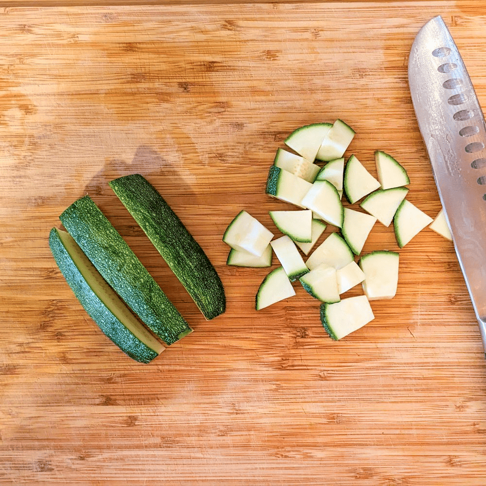 halved zucchini cut into thirds next to large diced pieces and a knife on a cutting board