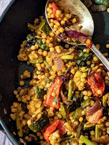 israeli couscous tossed in romesco pasta sauce and roasted vegetables in nonstick pan with a metal spoon