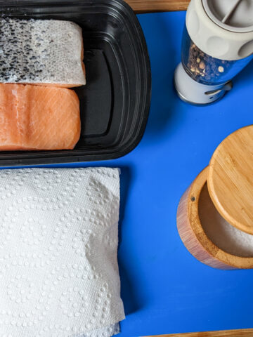 two salmon fillets in a shallow container next to a paper towel, pepper grinder, salt cellar on a plastic cutting board.