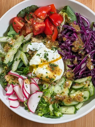 tuna avocado salad topped with a poached egg that's split open with yolk dripping out surrounded by diced tomatoes, shredded red cabbage, sliced cucumbers, sliced radishes, sliced celery on a bed of mixed greens.