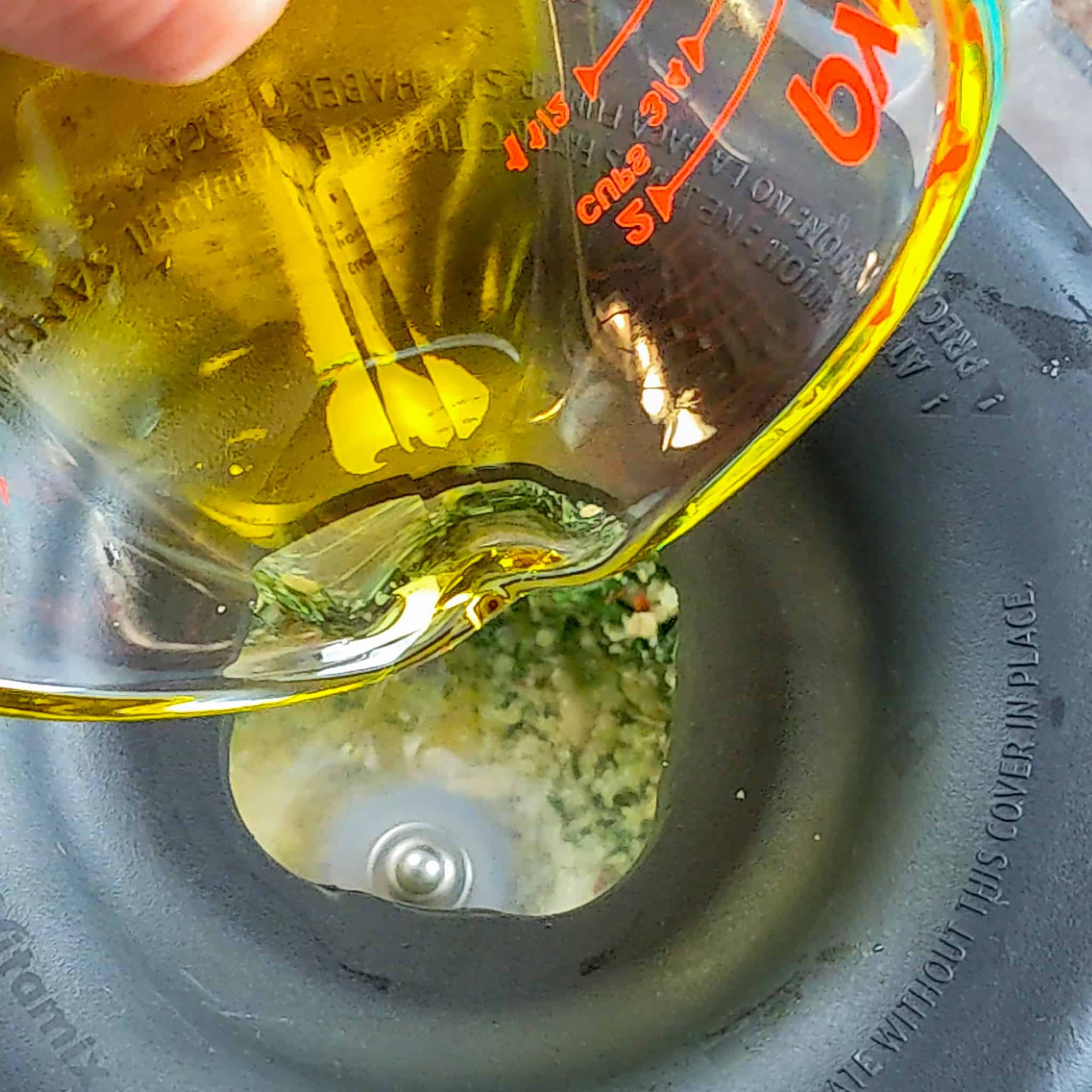 spicy basil pesto in a blender with oil in a glass measuring cup being poured through the blender lid's hole