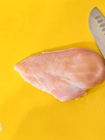 a boneless skinless chicken breast and a knife laying on a color coded plastic cutting board for raw chicken
