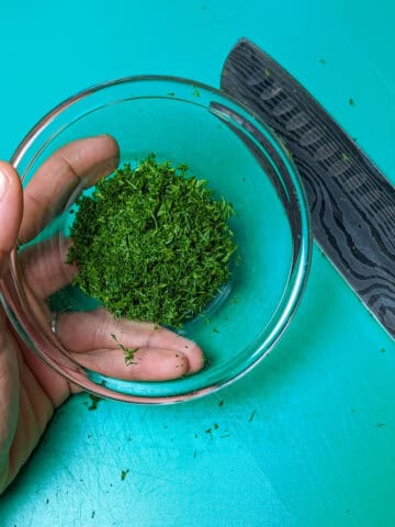 a hand holding a glass bowl of chopped dill over a plastic cutting board with a knife