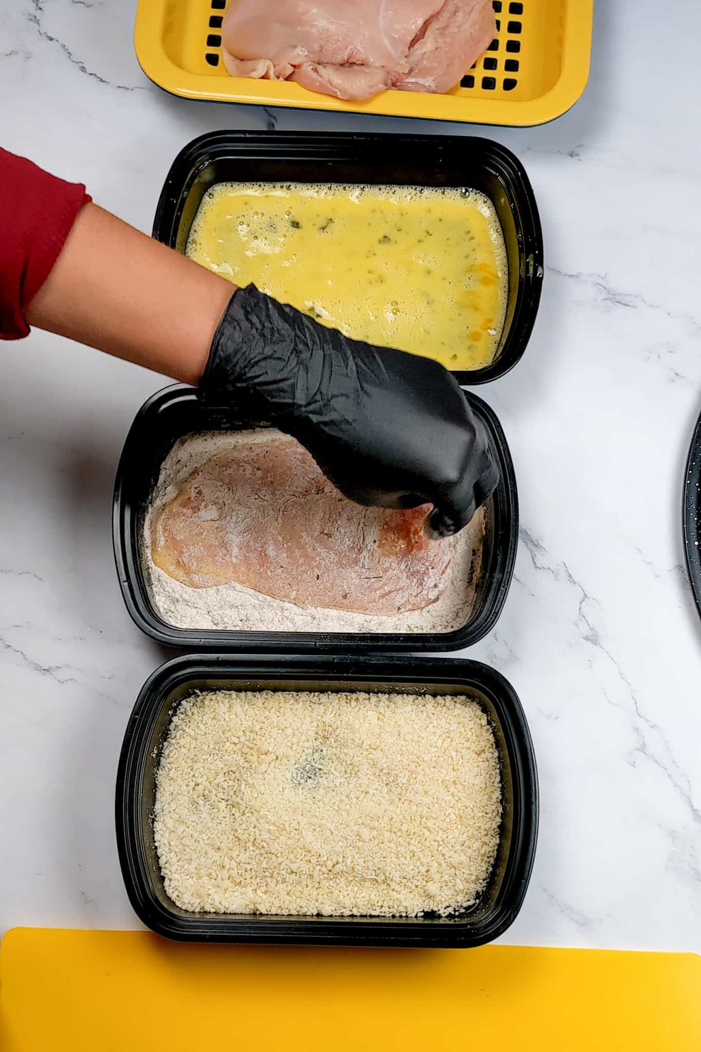 chicken cutlet being dredged in a shallow pan of flour assembled between two other shallow trays, one with beaten eggs and the other with bread crumbs