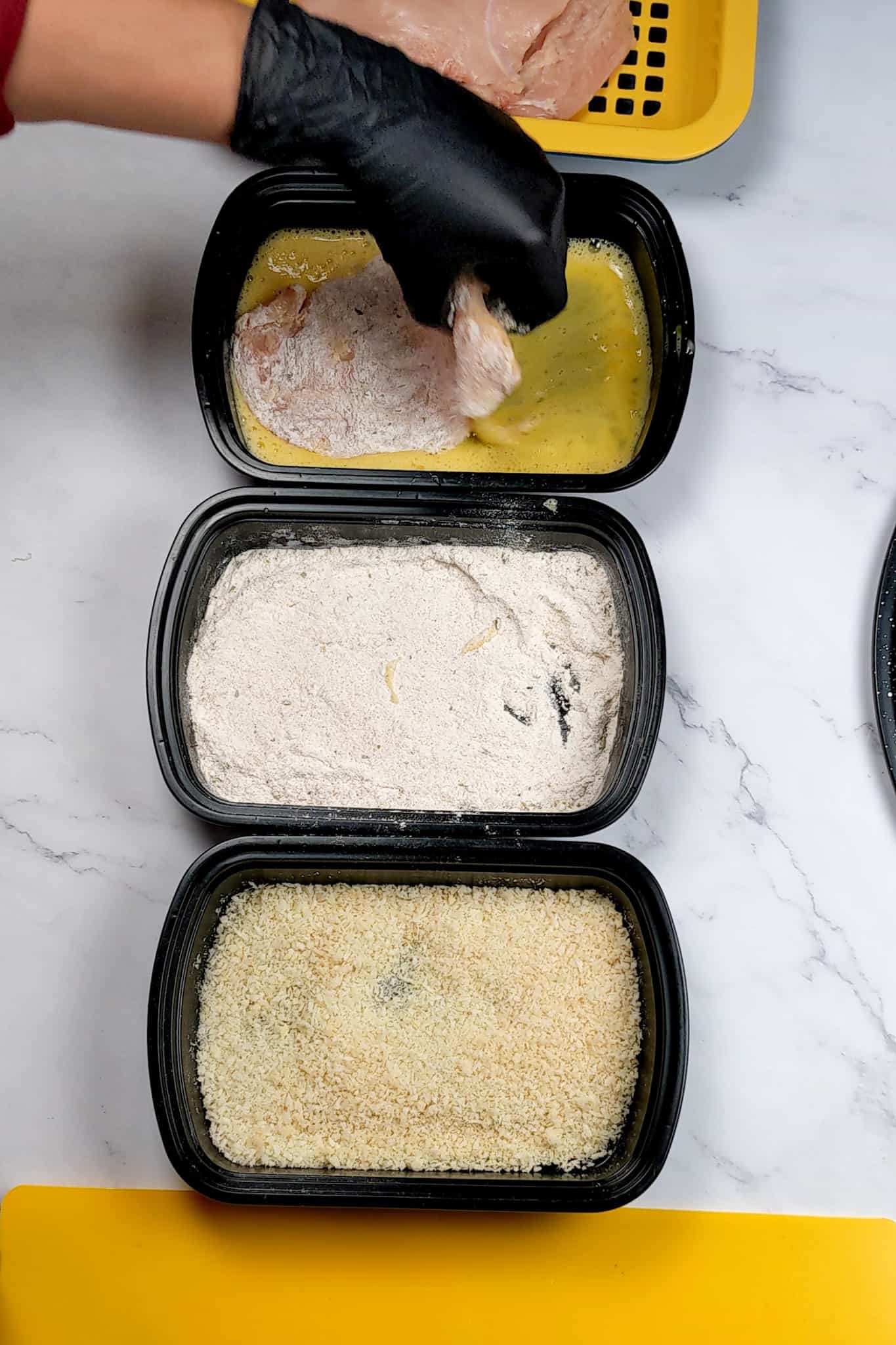 flour coasted chicken cutlet being dipped in a shallow pan of egg wash assembled next to other shallow trays, one with flour and the other with bread crumbs