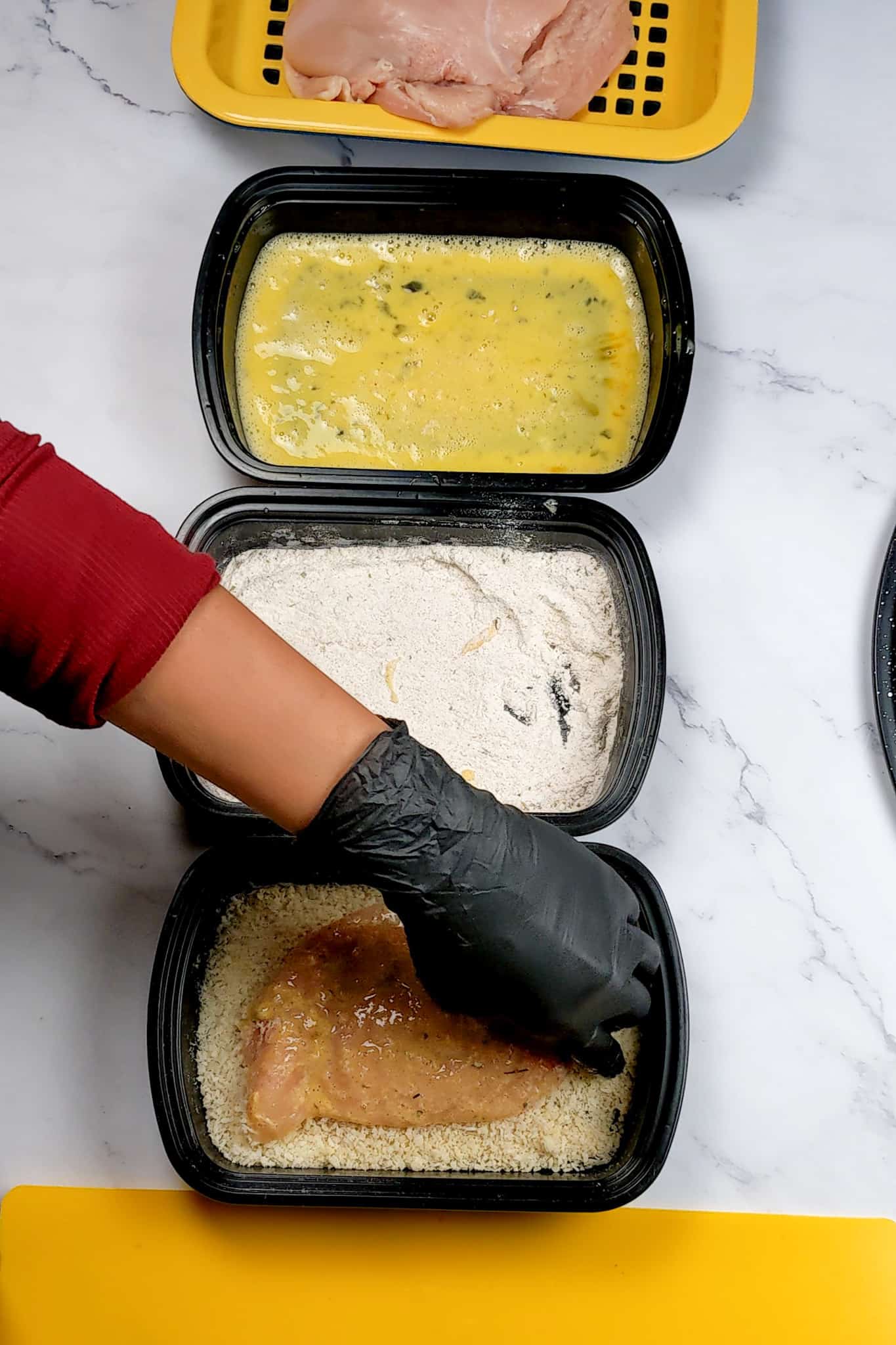 raw chicken cutlet being placed in a shallow pan of panko breadcrumbs assembled next to other shallow trays, one with flour and the other with beaten eggs