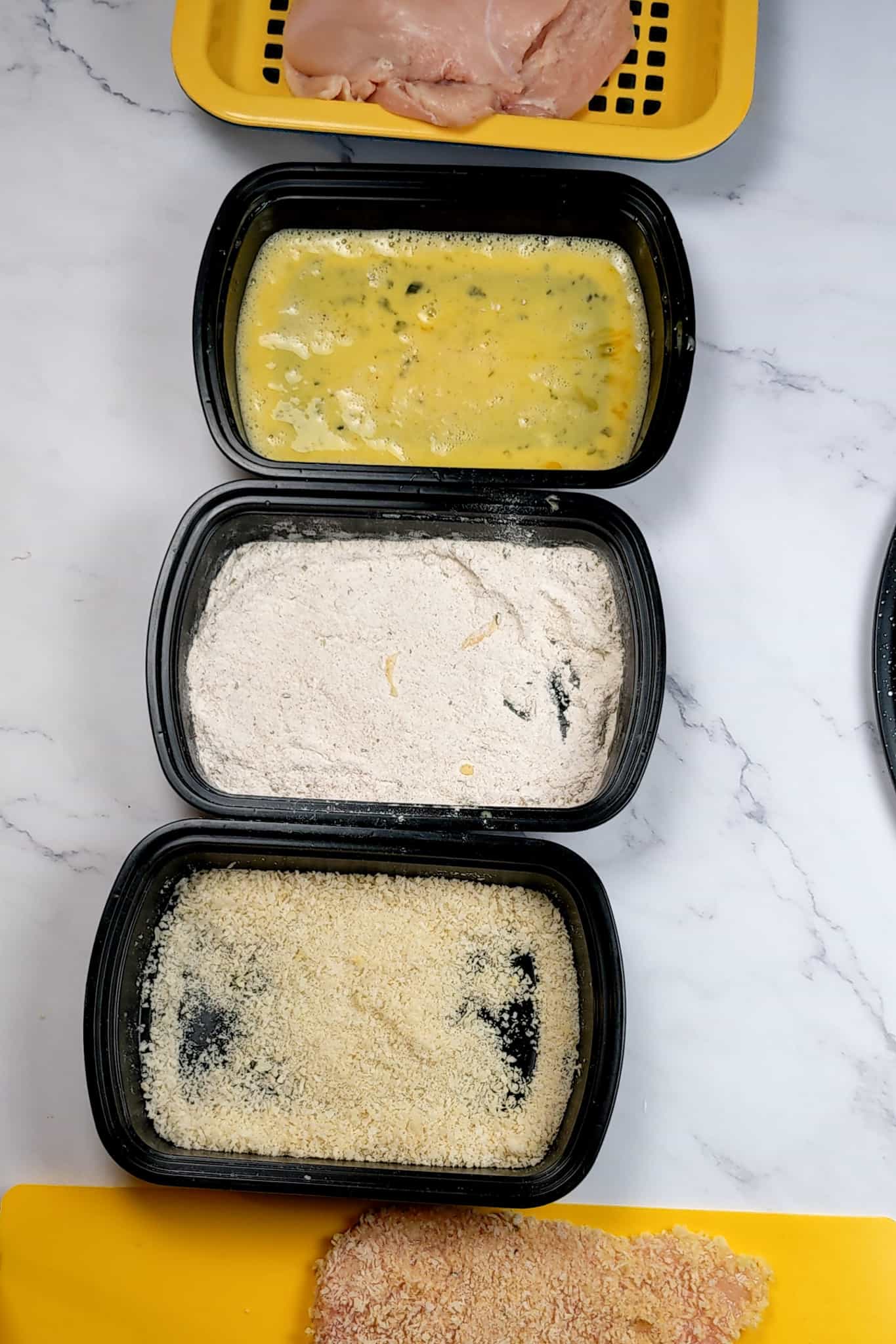 meat draining container with raw chicken cutlets assembled next to three shallow trays, one with beaten eggs, another with flour and the last one with panko breadcrumbs and next to that a yellow cutting board mat with a breaded raw chicken cutlet