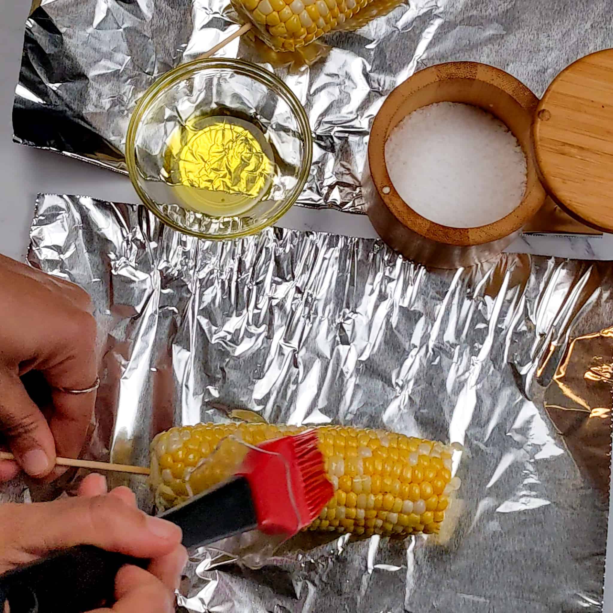 brushing olive oil on the corn with a pastry brush on aluminum foil surrounded by a salt cellar and a bowl of olive oil