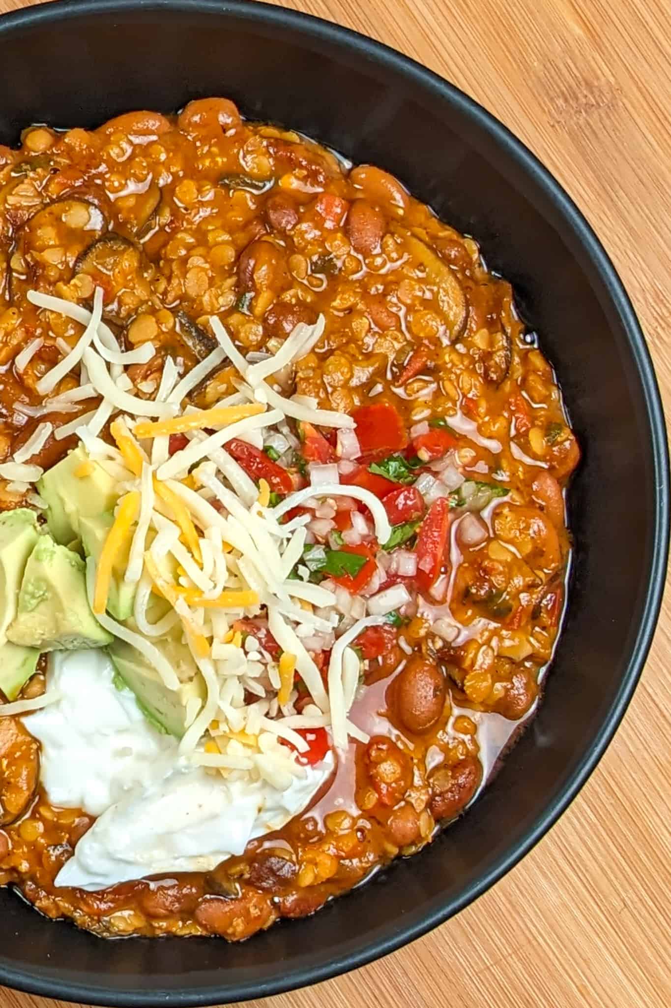 top view of Vegetarian Chipotle Mushroom Lentil Bean Chili in a wide soup bowl topped with sour cream, diced avocado, pico de gallo and pepper jack shredded cheese blend