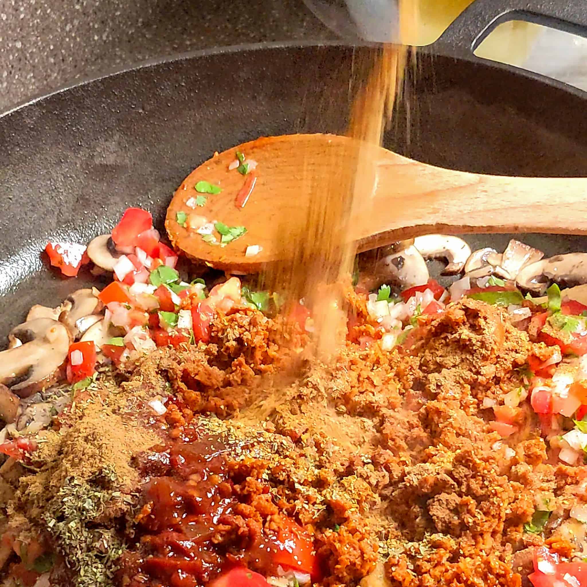 seasoning being added to the chipotle in adobo, soy chorizo, pico de gallo vegetables, and browned sliced mushrooms mixture in a large wide cast iron skillet