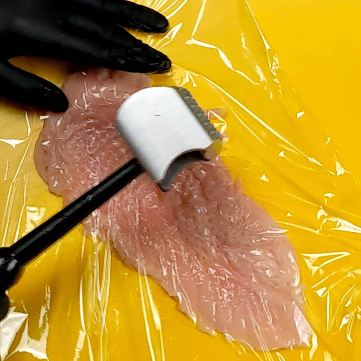 halved chicken being pounded out with a meat mallet on a yellow cutting board