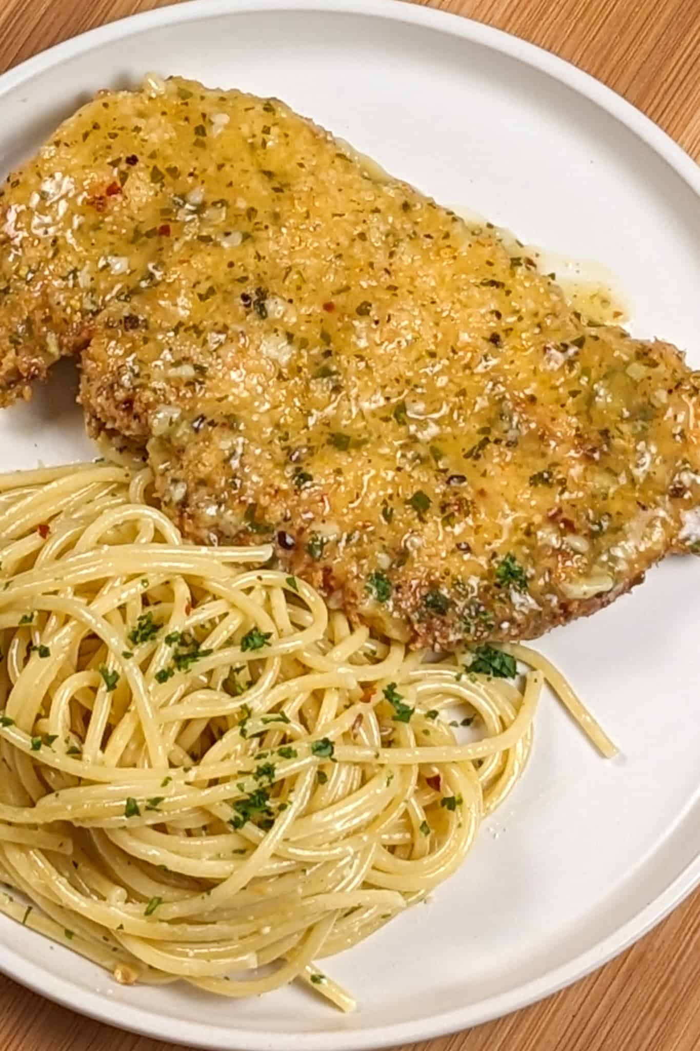 wet lemon pepper chicken cutlet on a white round plate next to olive oil and grated cheese tossed spaghetti dusted with chopped fresh parsley