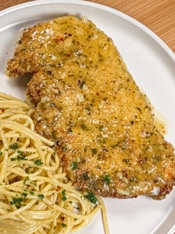 wet lemon pepper chicken cutlet on a white round plate next to olive oil and grated cheese tossed spaghetti dusted with chopped fresh parsley