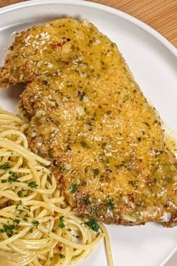 wet lemon pepper chicken cutlet on a white round plate next to olive oil and grated cheese tossed spaghetti dusted with chopped fresh parsley