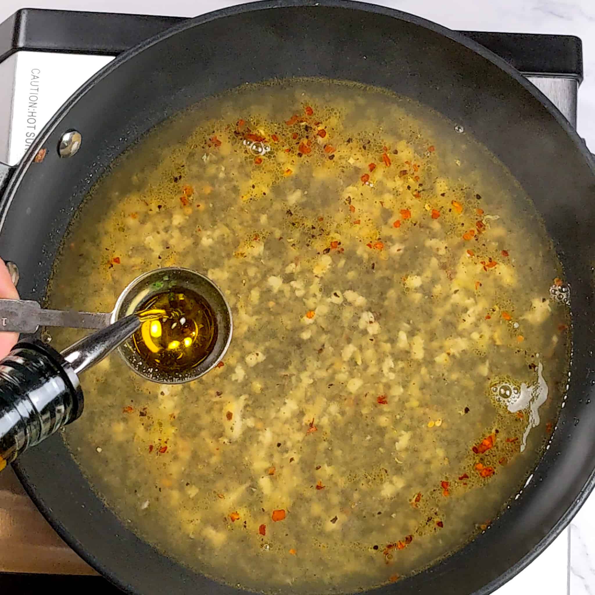 olive oil being poured in a 1 tablespoon measuring spoon over the unfinished lemon pepper sauce