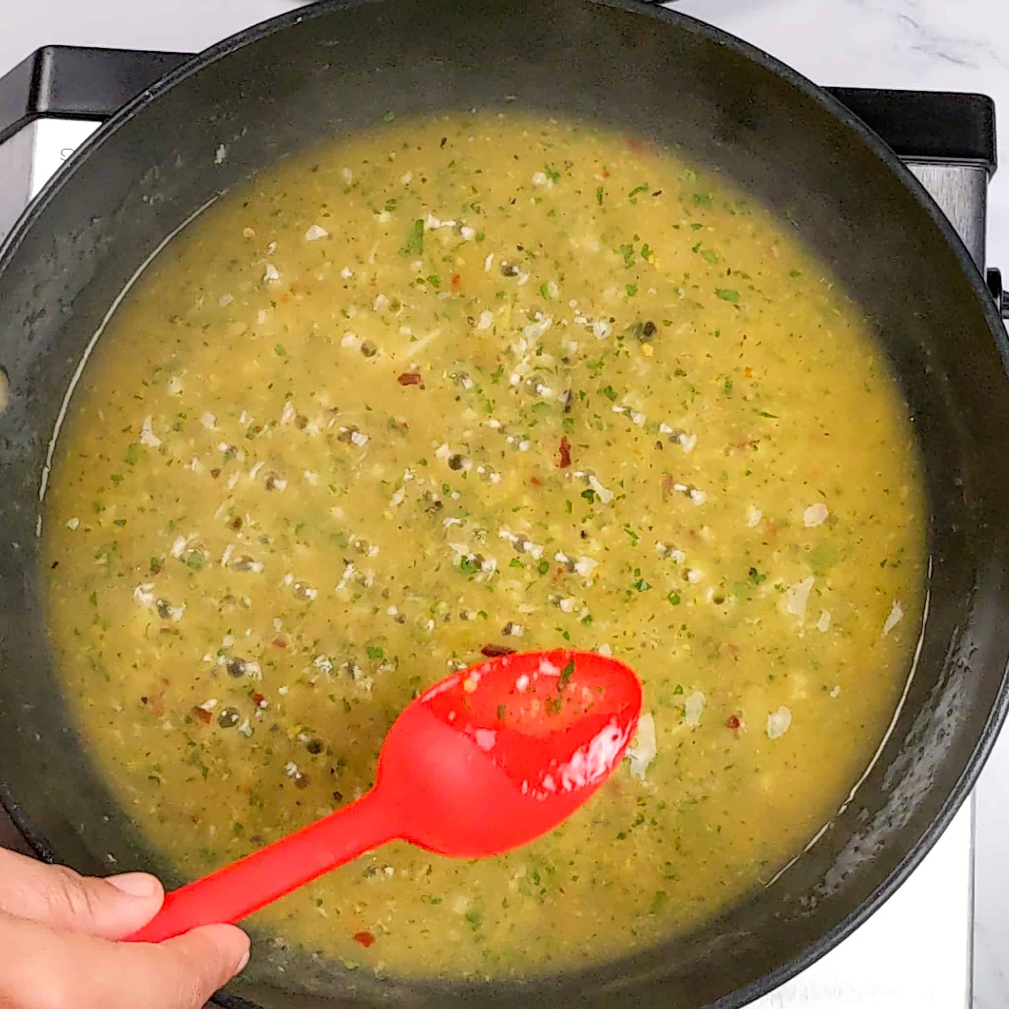 the wet lemon pepper sauce reducing with small bubbles in a non-stick frying pan being tasted with a silicone red spoon