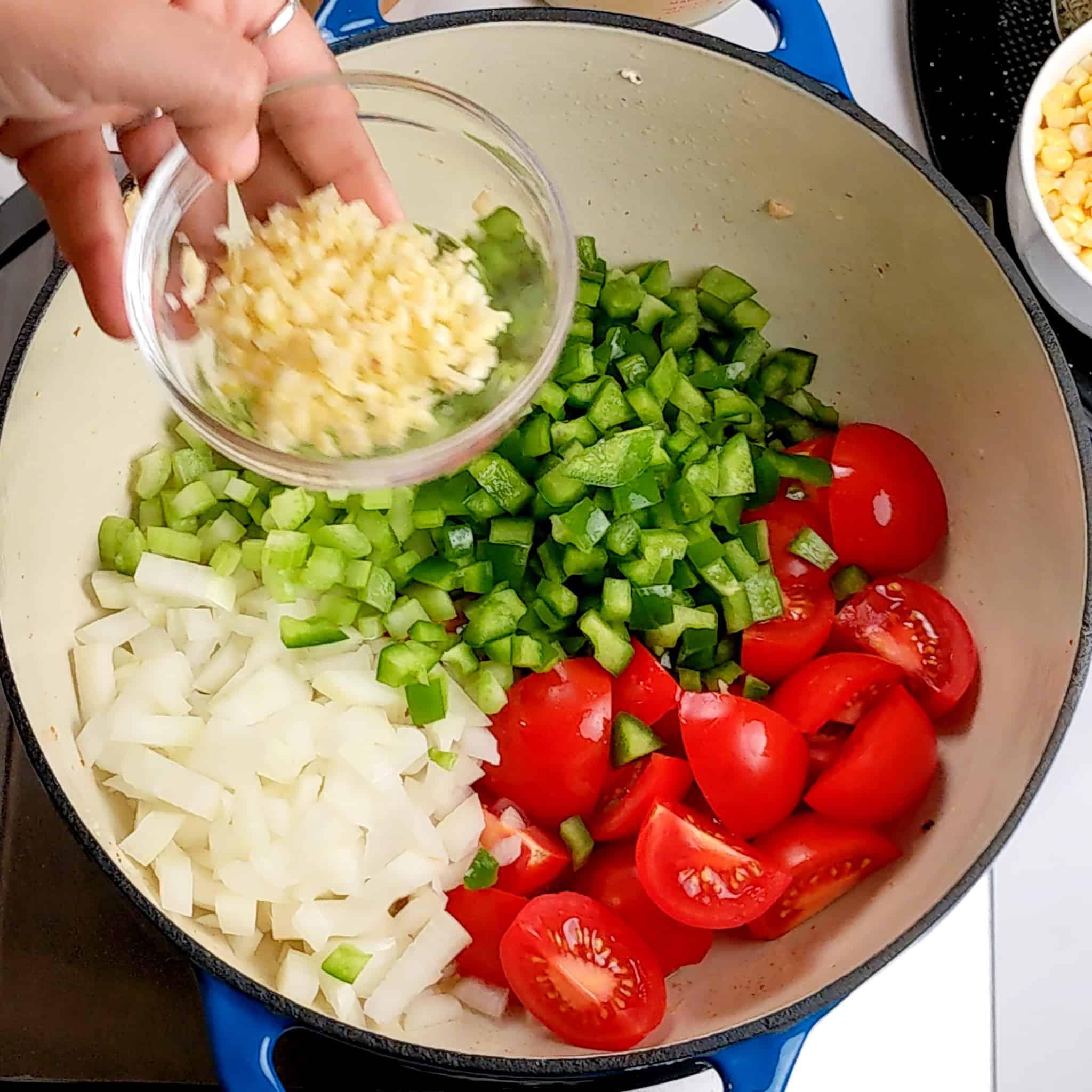 chopped garlic in a small glass bowl held over a dutch oven of quartered campari tomatoes, diced onions, green bell pepper, celery for the spicy calabraian chicken chowder