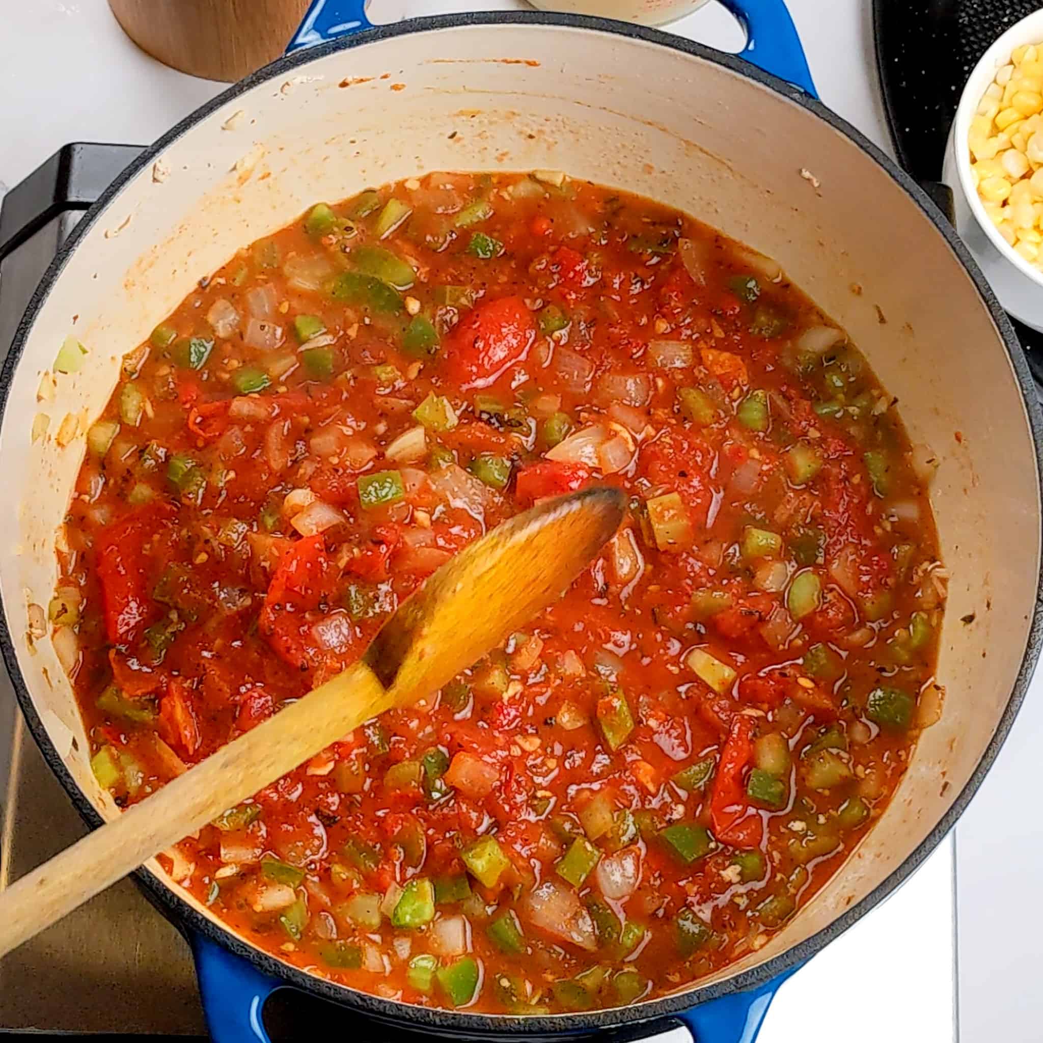 diced aromatic vegetables mixed with crushed fire-roasted tomatoes and crushed calabrian peppers mixed in a dutch oven with a wooden spoon laying in the pot