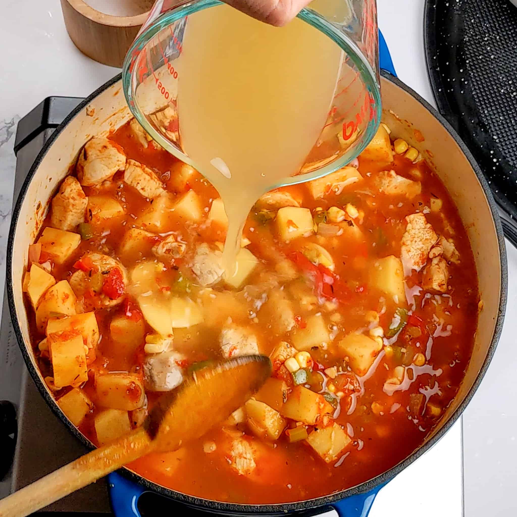 chicken broth being poured from a glass measuring cup into the spicy calabrian pepper chicken chowder in a dutch oven with a wooden spoon