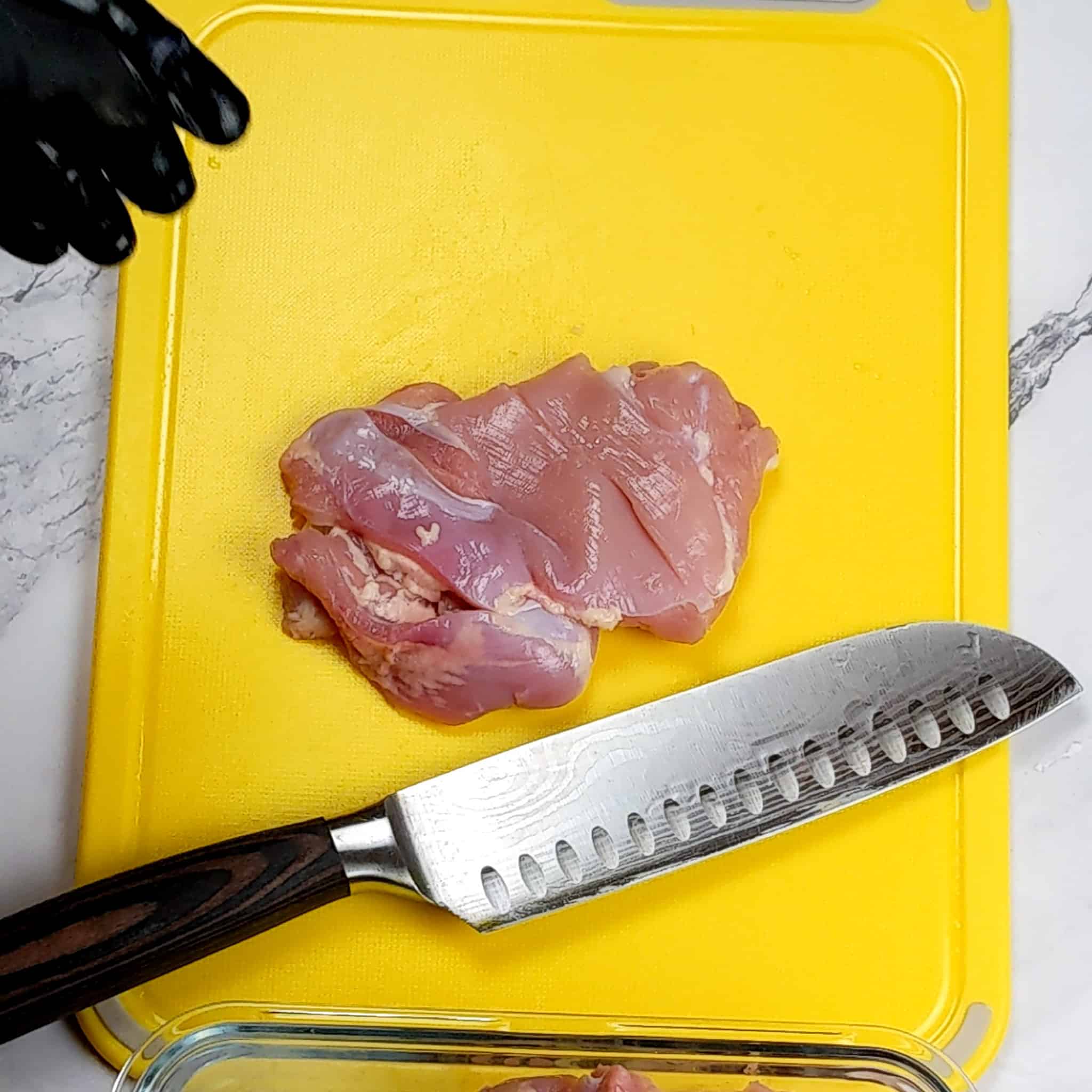 a chicken thigh on a plastic cutting board laying next to a santoku knife with three slits.
