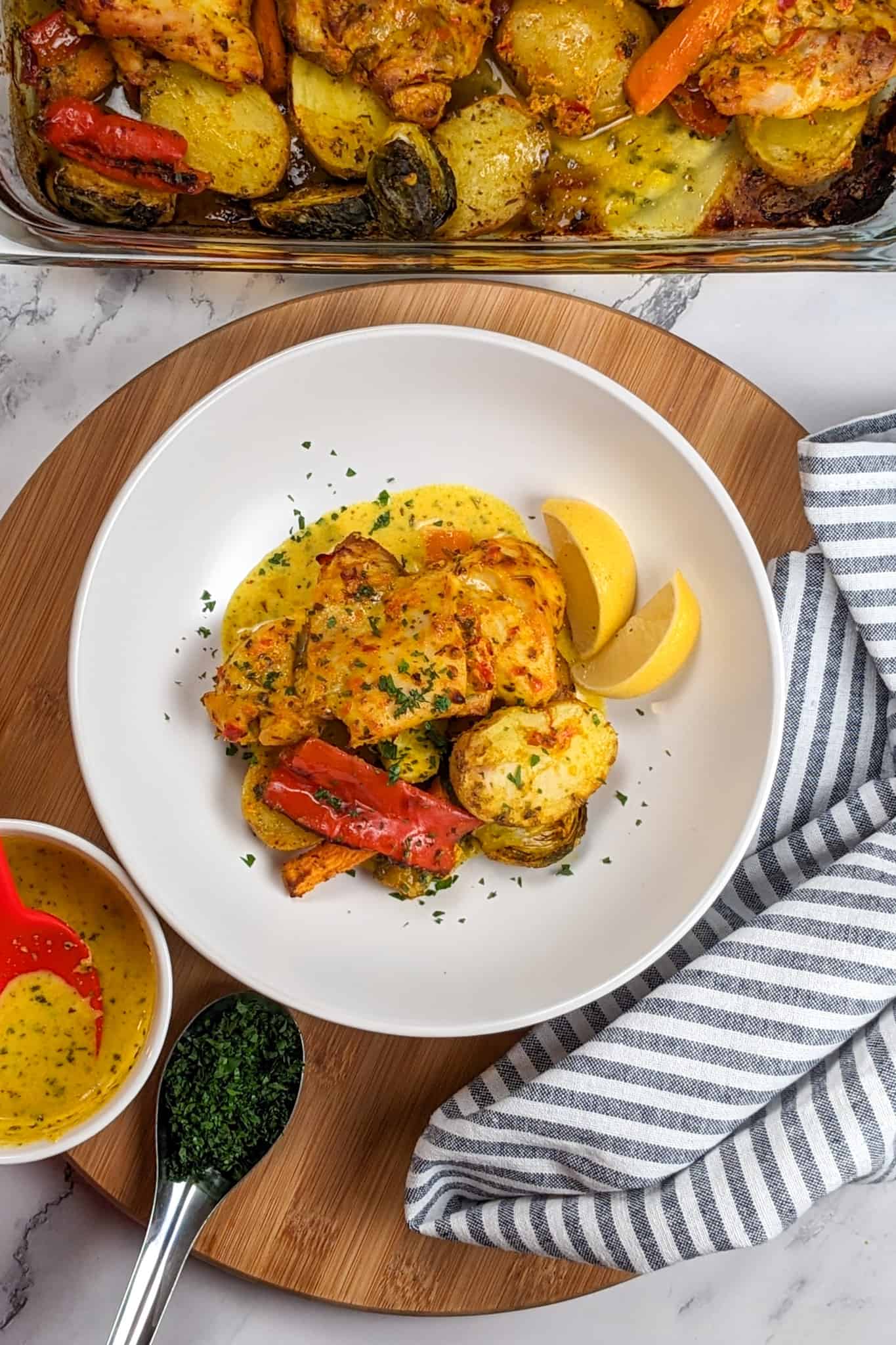 Top view the of Calabrian Pepper & Lemon Yogurt Baked Chicken and Vegetables dish next to a kitchen towel, the baked dish in a baking dish, yogurt sauce in a bowl and a spoon filled with chopped parsley.