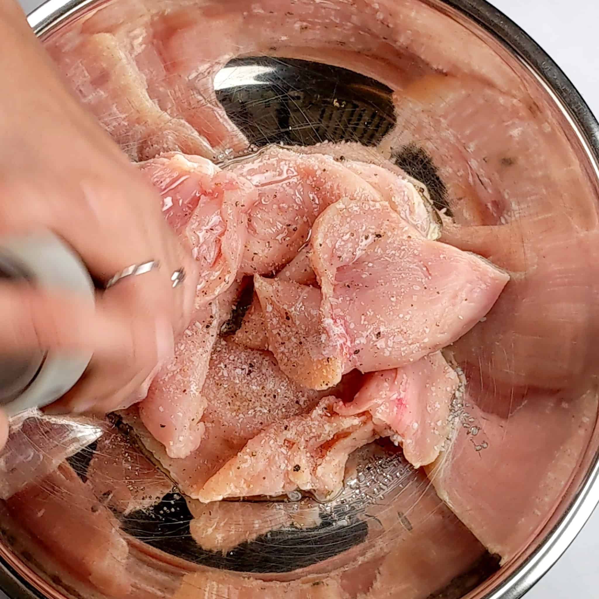 butterflied chicken breast seasoned with salt and pepper in a large stainless steel mixing bowl with black pepper being ground over it