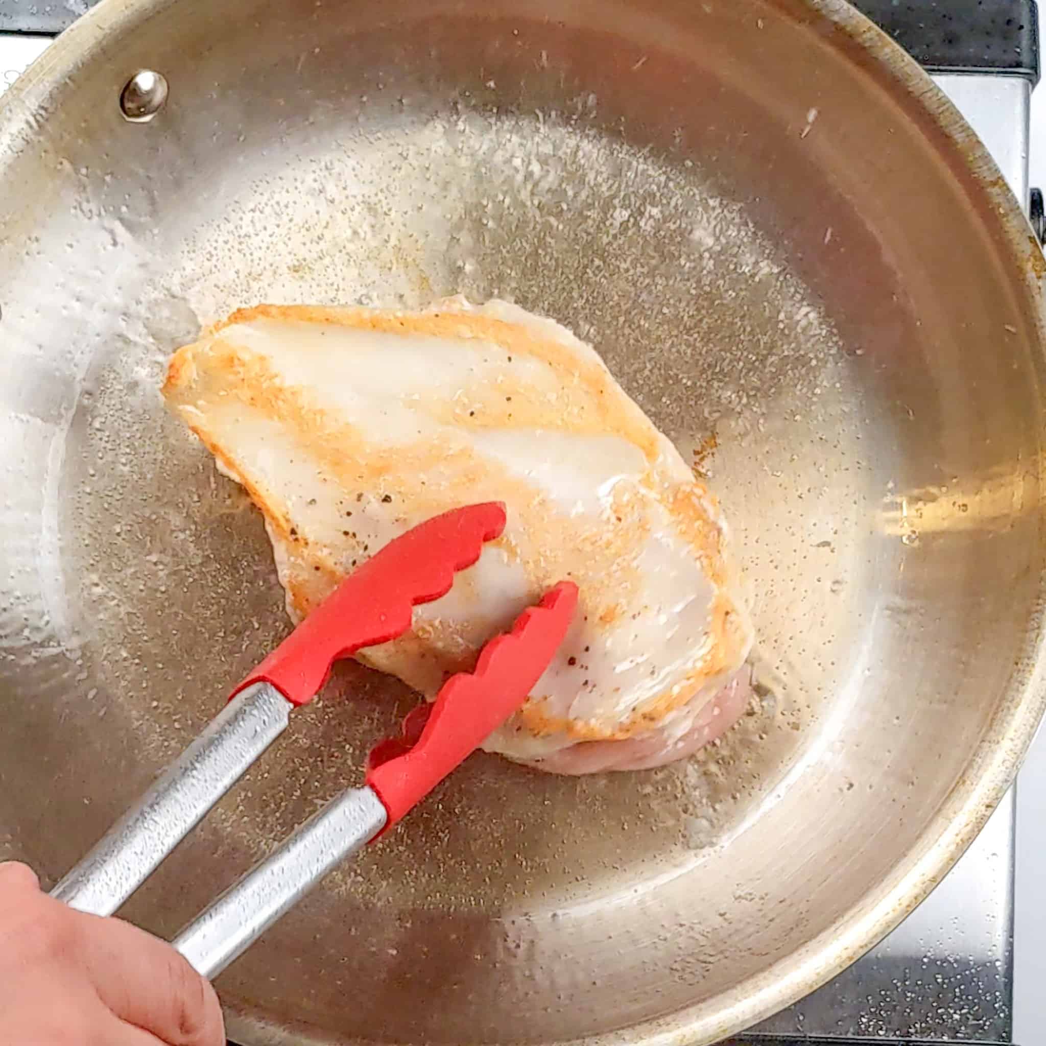 golden brown seared seasoned chicken breast in a stainless steel frying pan with oil being touched with a pair of KitchenAid silicone tongs