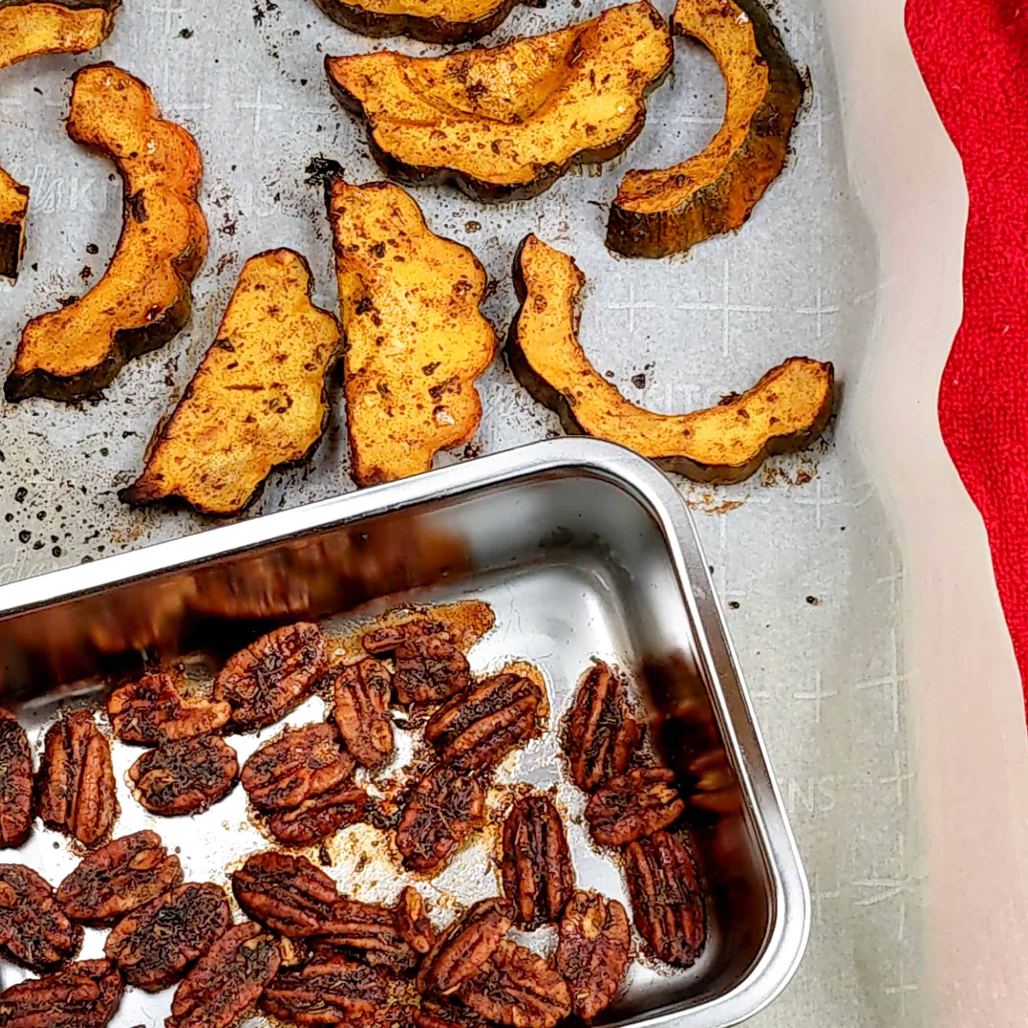 roasted sliced acorn squash and a small deep metal tray of maple syrup sauce coated pecans on a sheet pan lined with parchment paper