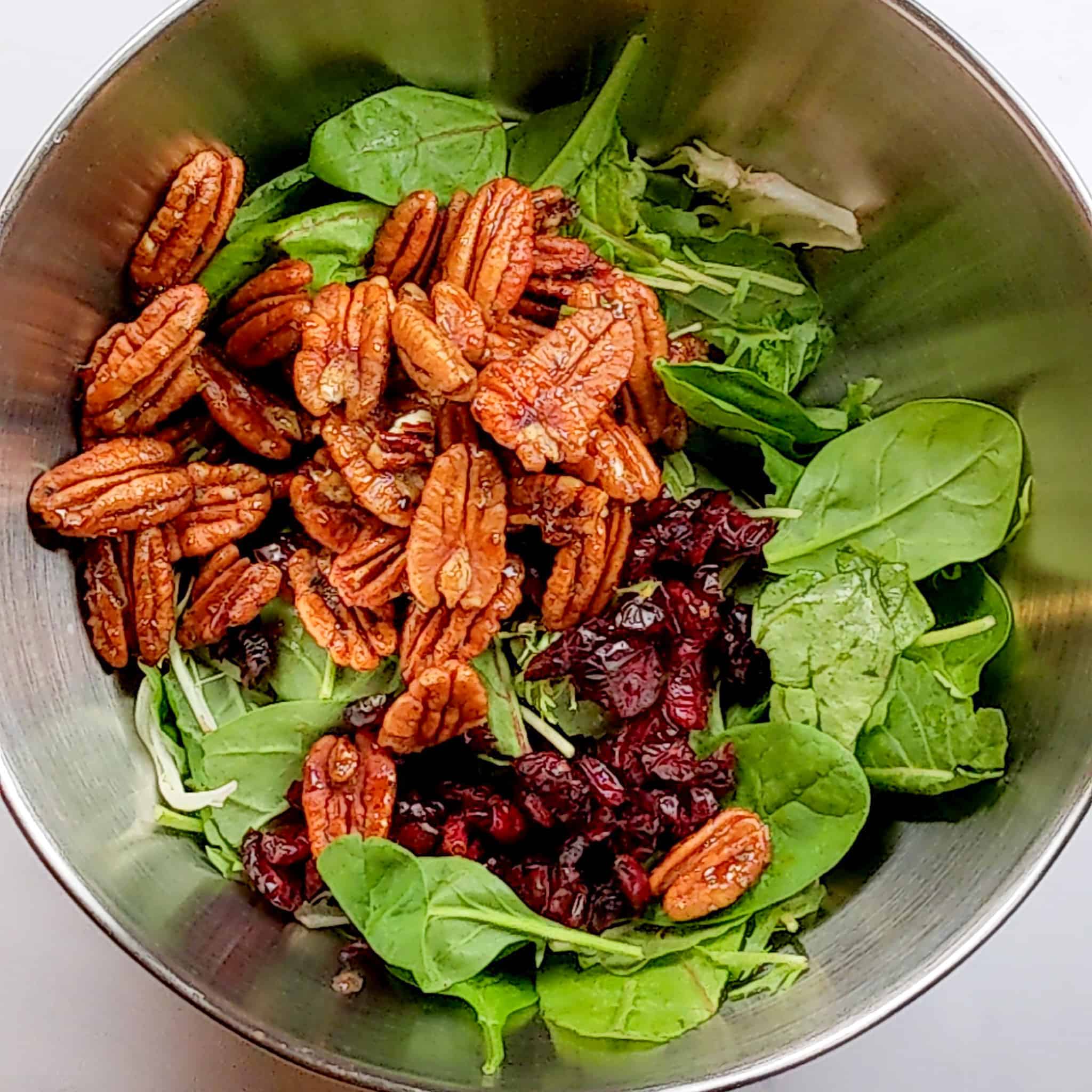 a stainless steel mixing bowl with mixed greens topped with candied pecans and dried cranberries.