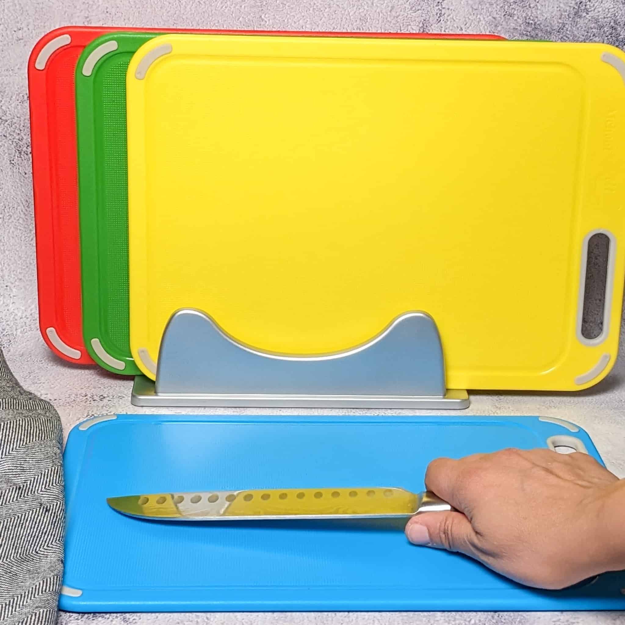 The BPA-Free Color Coded Plastic Cutting Board displayed with the fish board on the counter with a knife being held over it while the other three boards for raw chicken, meat and cooked foods are on it's stand up rack in the background