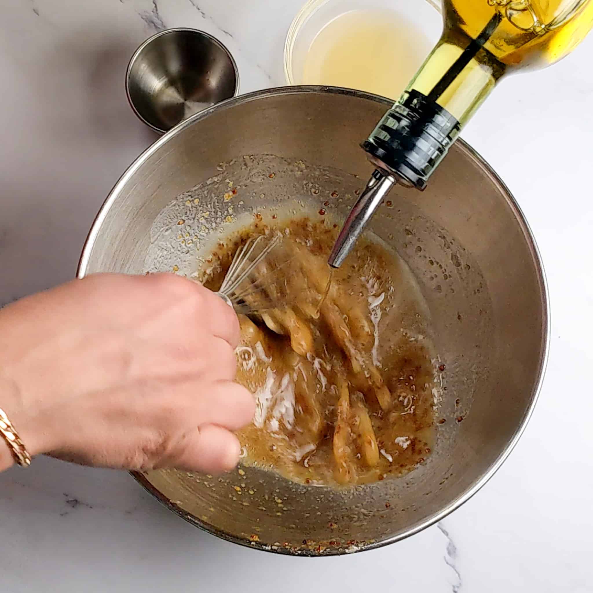 a hand whisking the dressing while pouring in oil into a stainless steel mixing bowl.