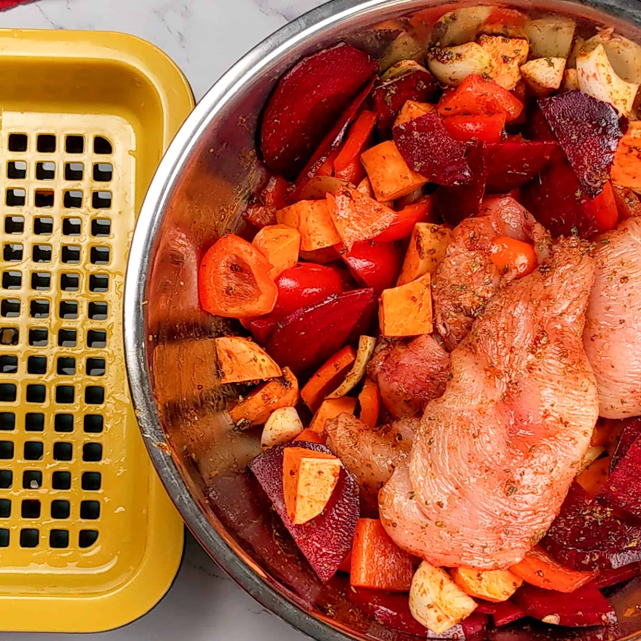 seasoned and tossed root vegetables with onion and butterflied chicken in a large wide stainless steel bowl next to a drainage tray