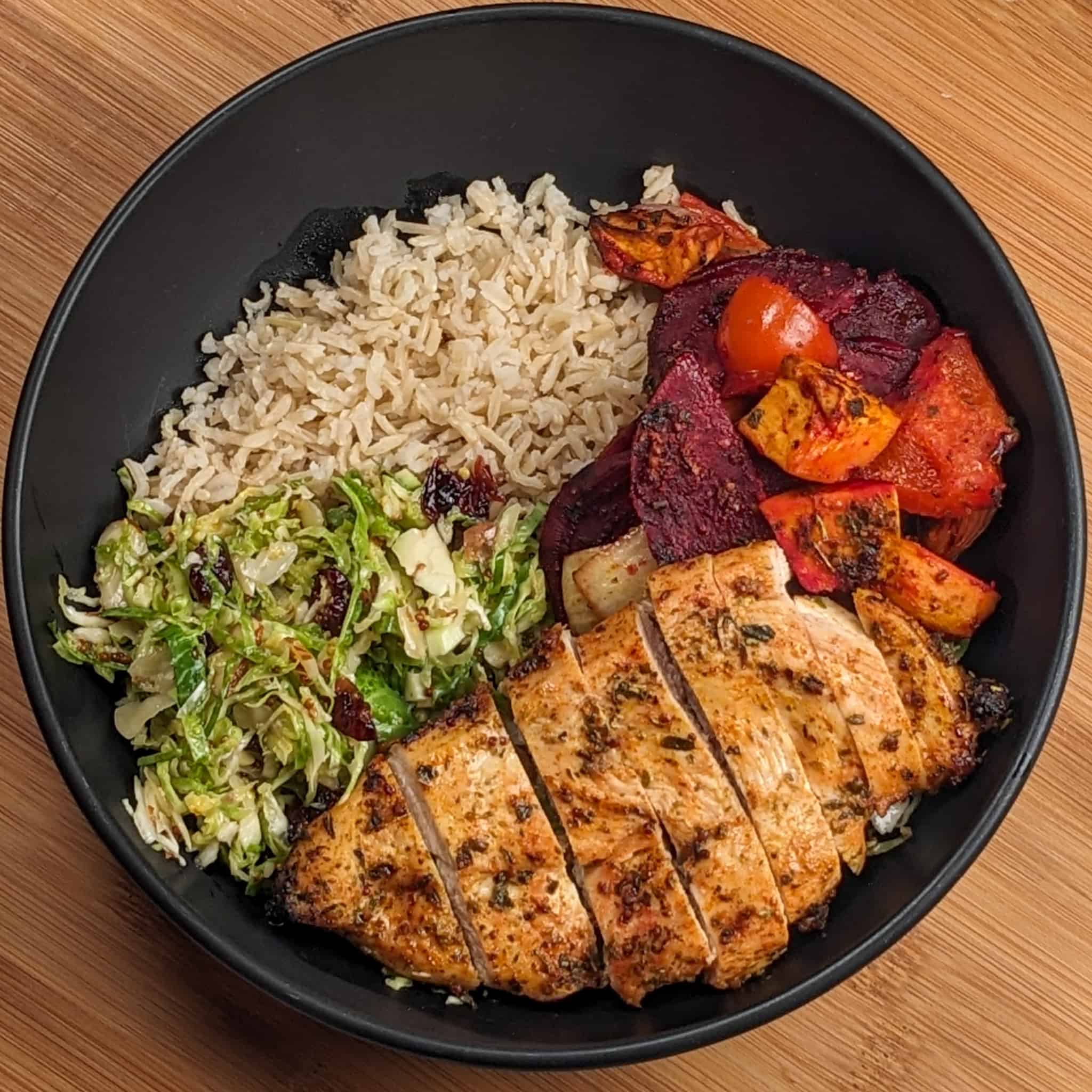 a wide rim bowl with brown basmati rice, root vegetables, sliced chicken breast and shaved brussels sprout salad